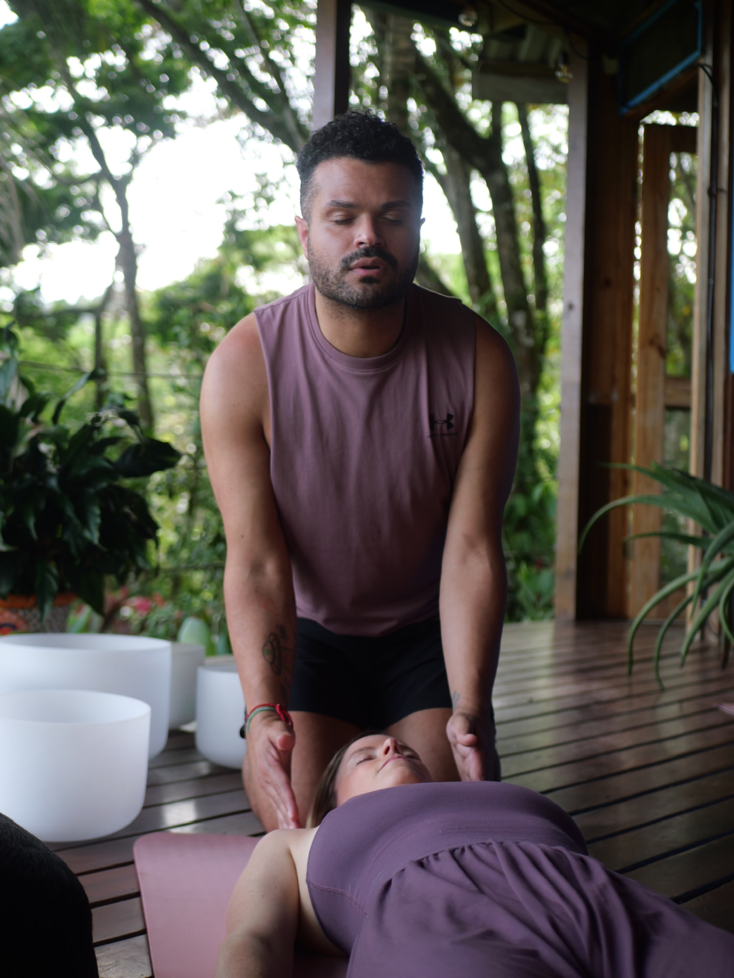 A man kneeling with closed eyes beside a woman resting on an open-air wooden deck surrounded by greenery